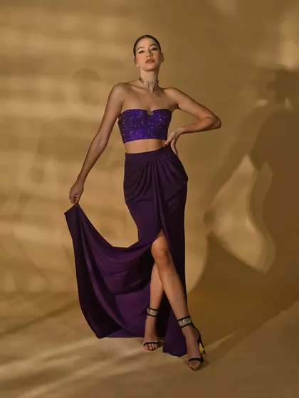 A shot from the 'Amethyst Edit' featuring a strapless top and draped skirt. The model's pose highlights the leg slit and the texture of the embellished top.