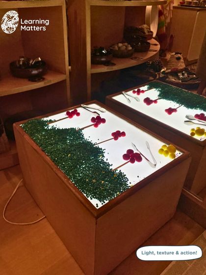 This light table is curated with colorful gems and sticks, inviting toddlers to explore light, texture, and fine motor skills. Using tongs to pick up the soft pompoms builds hand-eye coordination and finger strength.