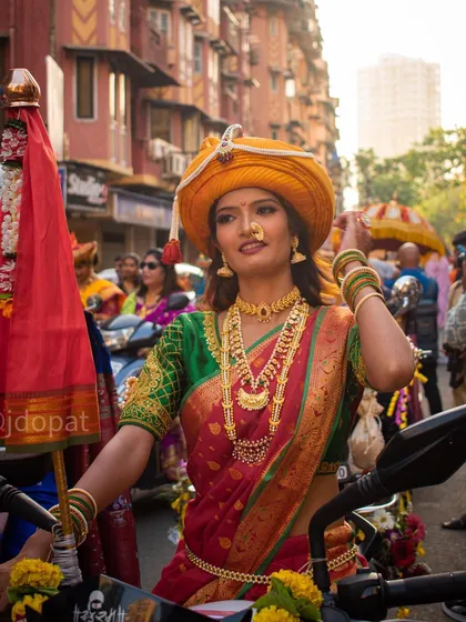 A woman in a beautiful red and green saree with a yellow turban holds a Gudi flag while sitting on a motorcycle during the festival.