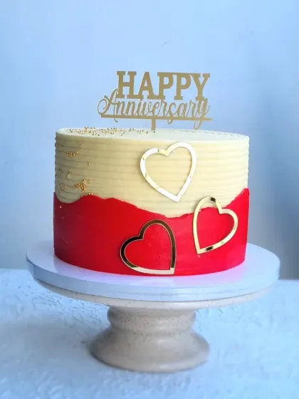 A modern anniversary cake with a red and white fault line design. The cake is decorated with gold leaf, a "Happy Anniversary" topper, and acrylic heart charms.