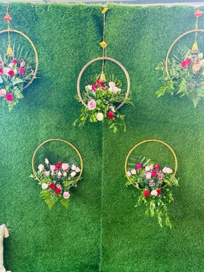 A simple and elegant Mehendi backdrop idea. This design uses an artificial grass wall decorated with five hanging golden hoops, each filled with beautiful arrangements of pink and white roses and green ferns.
