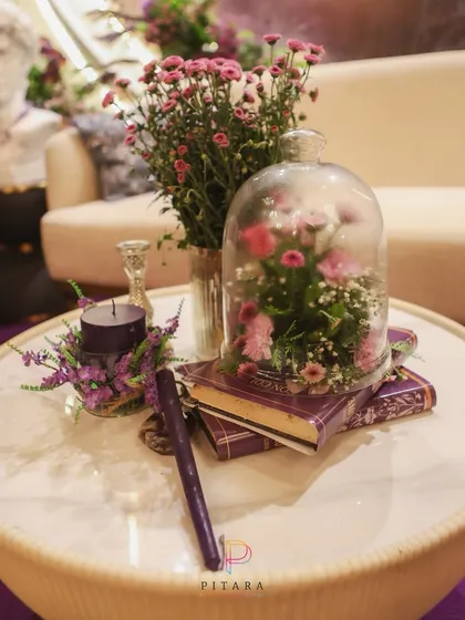 A romantic detail frozen in time. A delicate floral arrangement under a glass cloche, placed on vintage books with a dark candle, creates a beautiful, storybook moment on the coffee table.