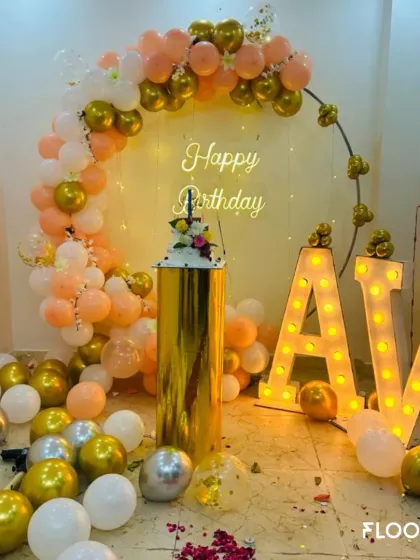 A chic and personalized birthday setup featuring a circular backdrop with a peach, white, and gold balloon garland. The design is completed with a 'Happy Birthday' neon sign, a gold pedestal for the cake, and large marquee letters for the initials 'AV'.