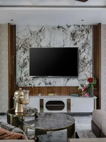 A composed and quiet lounge from our Chennai project. The TV is set against a dramatic marble wall, flanked by custom wooden shelving with brass details. A large sectional sofa provides ample seating.