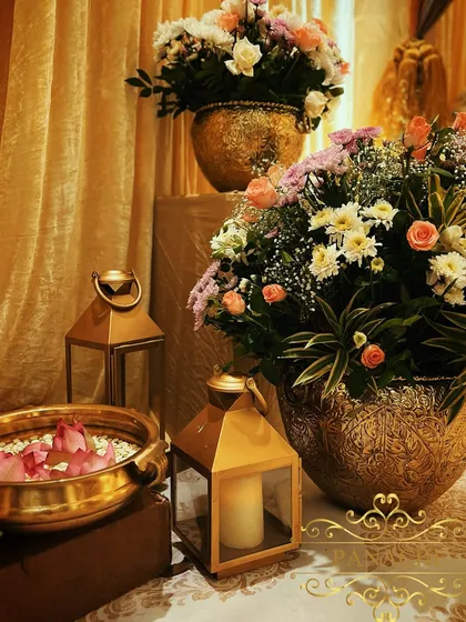 We use a combination of elements to create a welcoming atmosphere. Here, large floral arrangements in ornate golden vases are paired with lanterns at the entrance of the Roka ceremony.