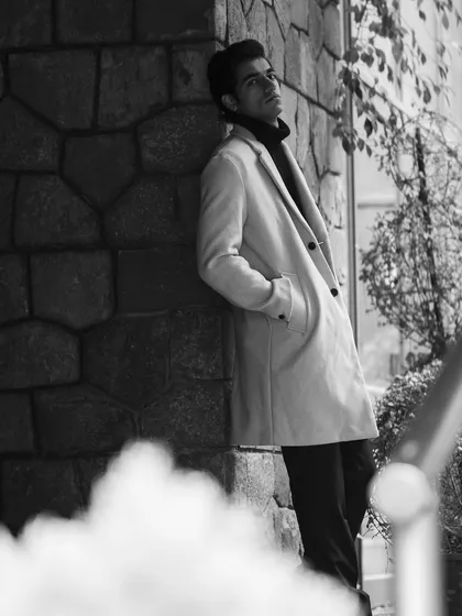 A model in a light-colored overcoat leaning against a stone wall. The composition and soft focus in the foreground create depth and a contemplative mood.