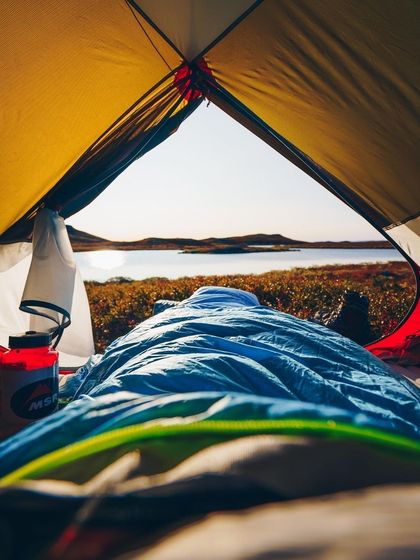 A first-person view from inside a sleeping bag, looking out over a tranquil lake at dawn. With a warm drink in hand, it’s the perfect way to start a day in the wild.