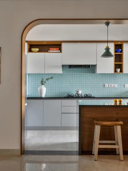 An arched entryway frames this kitchen, which features an island with a wooden base and bar stools. The soft blue-green tiles and white cabinets create a bright and welcoming space.