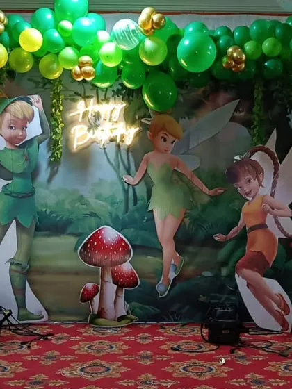 A whimsical Peter Pan and Tinkerbell theme. The setup features character cutouts against a forest backdrop with green and gold balloons, creating an enchanting Neverland experience.