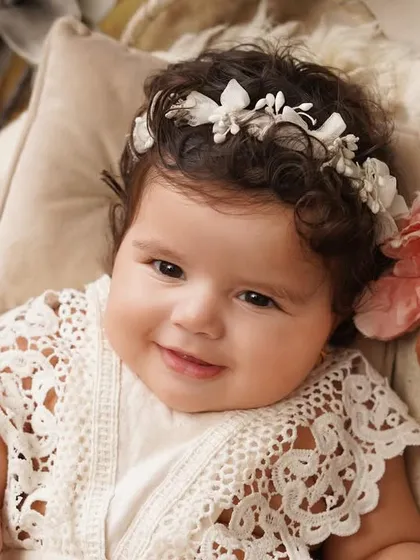 A heart-melting close-up of a smiling baby girl wearing a delicate lace outfit and a floral headband, looking directly at the camera.