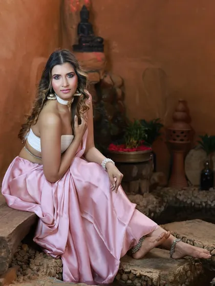 A full-length seated portrait in the rustic Aura Kalari setting. The pose is elegant and showcases the entire pink saree and white tube top ensemble.
