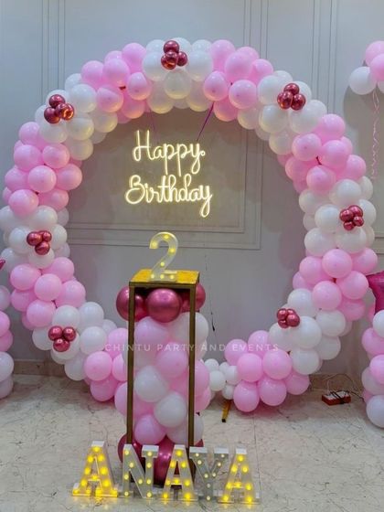 A pink and white balloon ring for a second birthday celebration. The setup is personalized with light-up marquee letters for the name 'Aanaya' and includes a pedestal for the cake.
