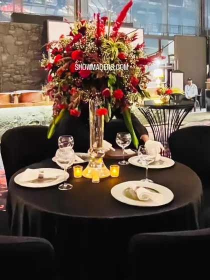 A close-up of a table setting for a dramatic evening event. The black tablecloth is contrasted with a stunning, tall centerpiece of deep red flowers, creating a bold and sophisticated look.