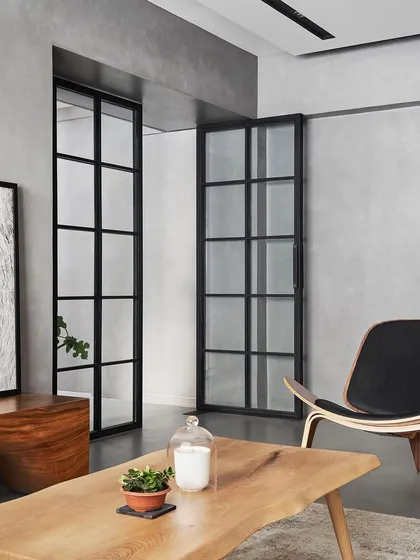 The living room features custom black-framed glass doors that lead to the kitchen, creating a sense of connection while maintaining distinct zones. The design uses a limited palette of concrete, white ash wood, and black steel to create a serene backdrop.