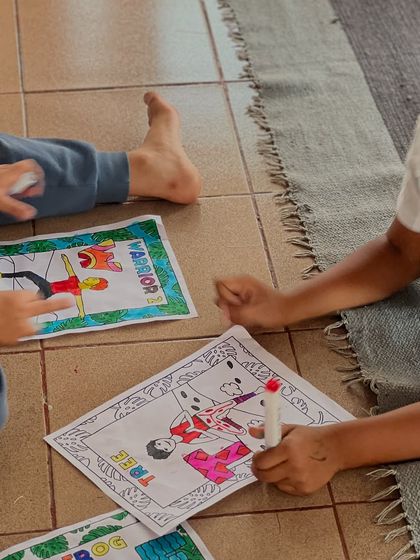 Children lie on the floor, focused on coloring sheets that depict various yoga poses like Warrior and Tree pose. This helps them learn the names and shapes of asanas in a fun way.