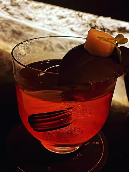 A dark red cocktail with a unique garnish: a thin chocolate disc and a small piece of candied fruit. This adds a dessert-like quality to the drink.