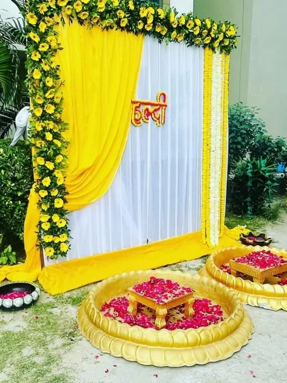 For an outdoor Haldi ceremony, I created this elegant setup featuring a frame draped in yellow fabric and decorated with yellow flower garlands. The arrangement includes traditional brass urlis and low stools for the rituals.