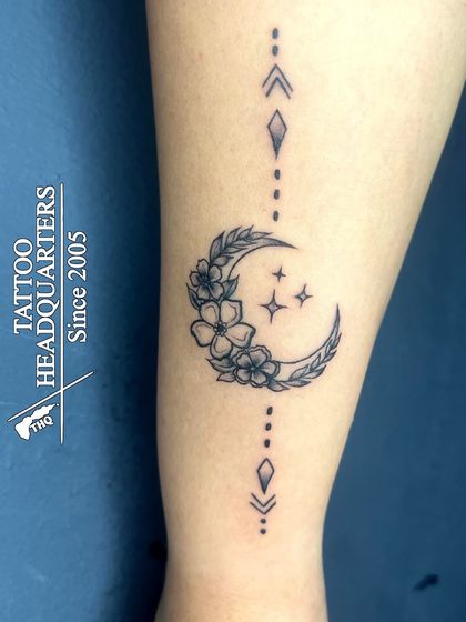 A beautiful crescent moon tattoo adorned with flowers and leaves, with geometric accents above and below.
