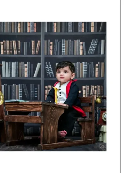 A full-length portrait of the baby wizard sitting at his desk. This shot captures the entire scene, from his bare feet to the towering bookshelves behind him.