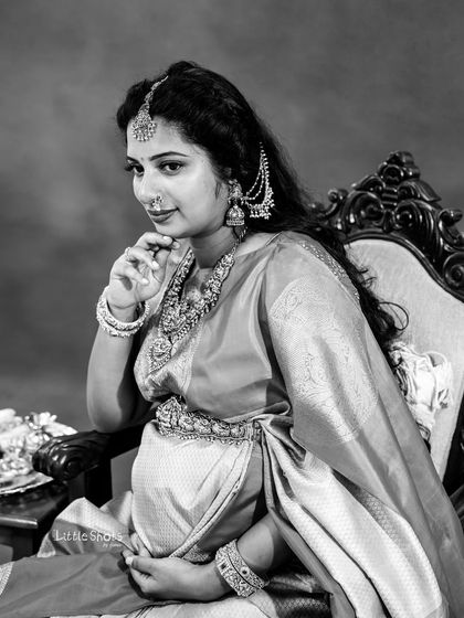 A classic black and white close-up of a mother-to-be in a traditional saree and jewelry. She is seated, looking thoughtfully to the side, creating a timeless and elegant image.