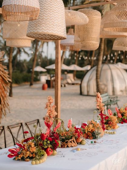A long table at the beach event, with hanging rattan lamps and a vibrant floral runner.