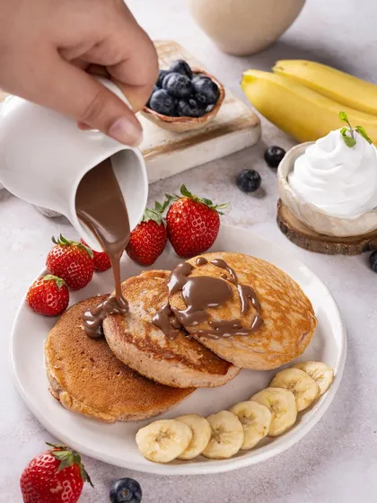 The moment of pouring chocolate sauce over a stack of fresh pancakes. This dynamic action shot is designed to be mouthwatering and visually satisfying.