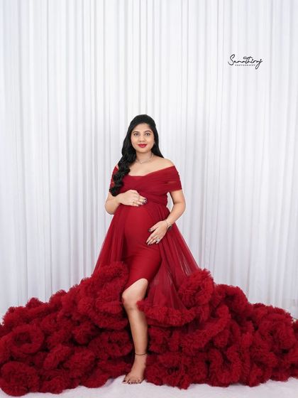 A full-length portrait of a mother-to-be in a dramatic red ruffled gown. The simple white background emphasizes the volume and color of the dress, creating a powerful statement.