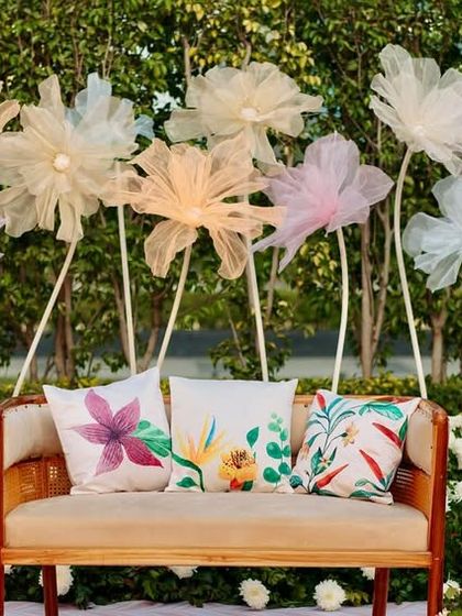 The Haldi seating stage, featuring a simple wooden bench with colorful printed cushions, set against a backdrop of lush greenery and whimsical fabric flowers.