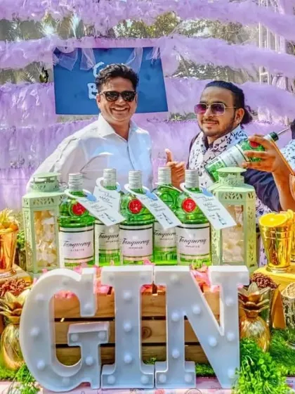 A happy guest and bartender pose behind the fully stocked gin bar. The setup features multiple bottles of Tanqueray gin, a large light-up 'GIN' sign, and an extensive selection of fresh garnishes for a vibrant party.