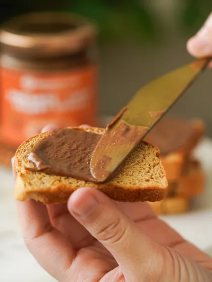 A close-up action shot of the chocolate spread being applied to a piece of toast, a simple yet powerful image of indulgence.