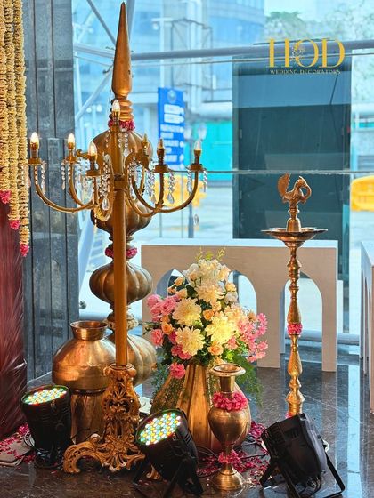 An elegant arrangement of traditional decor items, including a tall brass candelabra and a 'Kuthu Vilakku' (lamp), beautifully lit and surrounded by flowers.