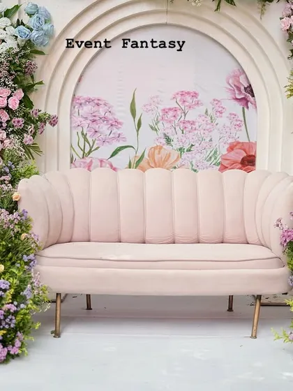 A closer look at a pastel-themed engagement stage. The blush pink scalloped sofa is framed by abundant arrangements of lavender, pink, and blue flowers, set against a backdrop with a delicate floral painting.