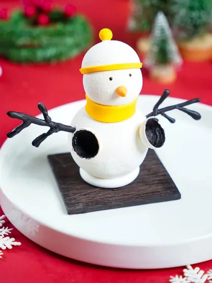 An adorable snowman dessert, presented simply on a white plate against a festive red background. This minimalist shot highlights the cute and playful character design.