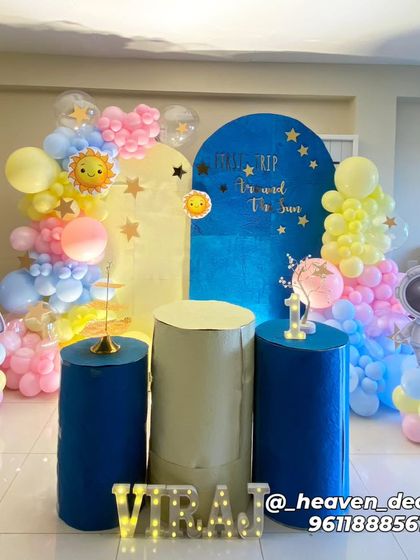 A "First Trip Around The Sun" space-themed birthday party. This cute setup features astronaut and rocket cutouts, a sun and stars backdrop, and a pastel-colored balloon garland.