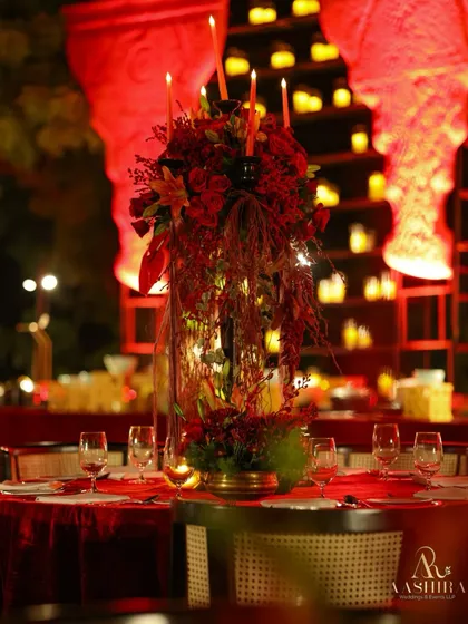 A tall, dramatic centerpiece of red roses and lilies on a dining table, with a backdrop of glowing candles and red-lit architectural elements.