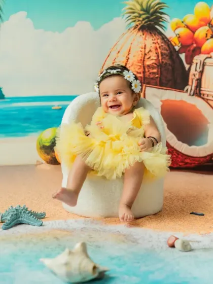 A joyful 4-month-old enjoying her 'Beach Baby' session. Dressed in a bright yellow outfit, she sits comfortably in a mini chair against a tropical backdrop, creating a perfect summer memory.