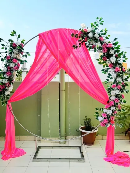 For smaller functions or outdoor events, I can provide a simple and elegant metal arch. This one is decorated with bright pink drapes, artificial flower arrangements, and fairy lights, serving as a beautiful and budget-friendly photo point.
