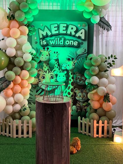 A "Wild One" jungle-themed first birthday for Meera. The setup features a dark green backdrop, cute animal cutouts, a wood-textured plinth, and a light-up number '1'.