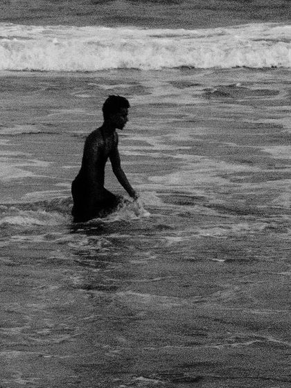 A boy wades into the ocean surf, his dark figure contrasting with the white waves in a classic black and white beach photograph.