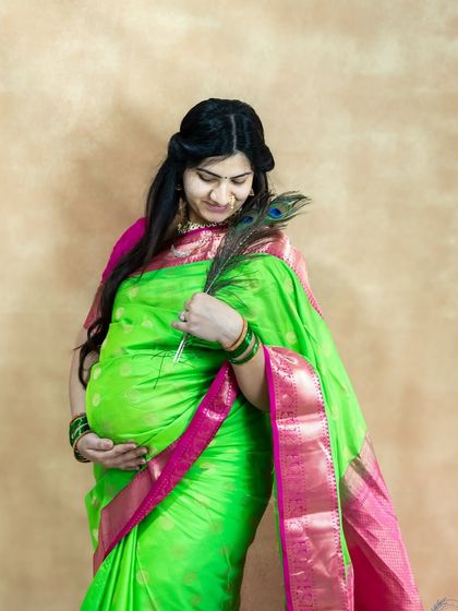 A lovely studio portrait of a mom-to-be in a green and pink saree, holding a peacock feather and looking at her bump.