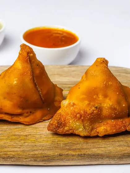 Two perfectly fried samosas on a wooden board, with dipping sauces and onions in the background. This classic Indian snack is shot on a white background, ideal for a clear and appealing menu listing.