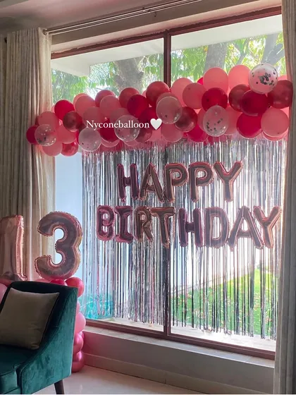 A pretty-in-pink theme for a 13th birthday. This home decoration features a silver fringe curtain, a garland of pink and red balloons, and large rose gold foil balloons for the number '13'.