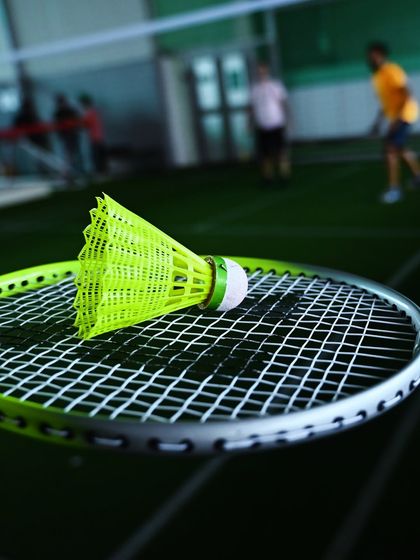 A close-up of a bright yellow shuttlecock resting on a badminton racket, with a game happening in the background. It’s these small details that make up the big moments on my courts.