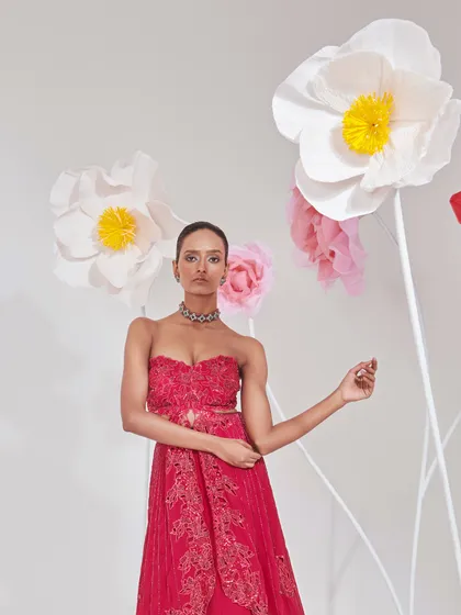 A full-length shot of a model in a red embroidered gown with a sweetheart neckline. The styling is clean and elegant, with the oversized paper flowers in the background adding a touch of whimsy and artistry to the scene.
