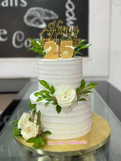 A classic and elegant two-tiered cake for a 25th anniversary. The pure white cake is adorned with fresh white roses, greenery, and a gold "Happy Anniversary" topper.