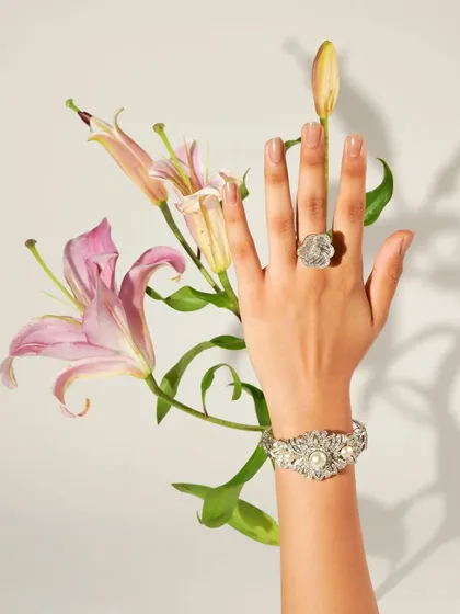 A model's hand, adorned with a floral-inspired ring and bracelet, is elegantly posed with fresh lilies. This image blends the beauty of nature with the intricate craftsmanship of fine jewellery.