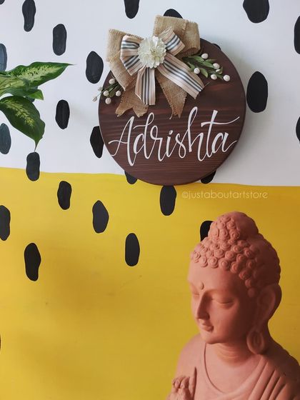 A round wooden name board for "Adrishta" styled in our studio. The combination of the dark wood, burlap bow, and handmade flowers creates a rustic yet elegant feel.