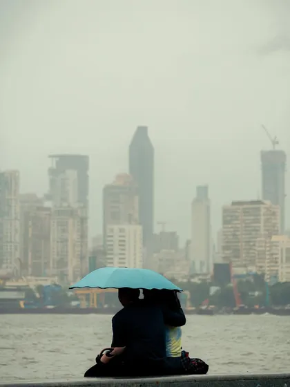 A couple sits by the sea under a bright blue umbrella, with the city skyline hazy in the distance. The image evokes a sense of shared intimacy and escape from the city.