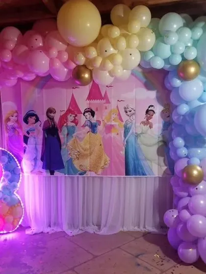 A princess castle theme for a third birthday party. The backdrop shows a castle with several princesses, framed by a pastel balloon arch and a large light-up number '3'.