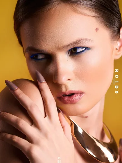 This cover for Roidx Magazine is a powerful beauty and accessory shot. The sharp lighting highlights the model's features, the bold blue eyeliner, and the smooth, sculptural gold neckpiece, creating a look of modern femininity.